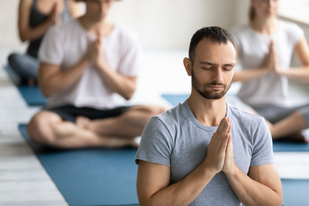 En man sitter i lotusställning på en yogamatta med händerna i böneposition och ögonen slutna, fler personer i bakgrunden.