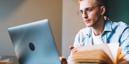 Ung man med glasögon studerar vid en bärbar dator med öppna böcker på ett bord.