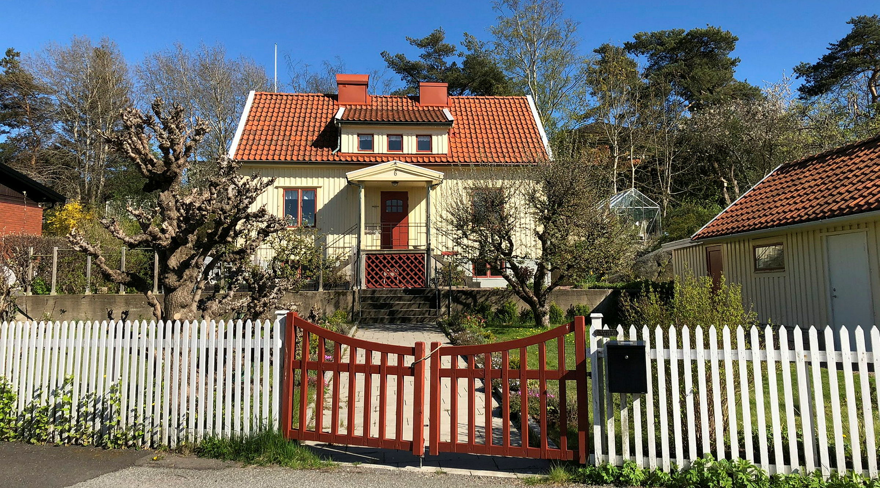 Ett gult trähus med rött tegeltak, vit staket och trädgård i soligt väder.