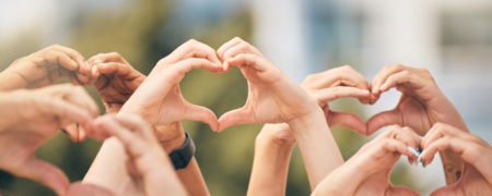 Flera händer formar hjärtan tillsammans mot en suddig bakgrund utomhus.