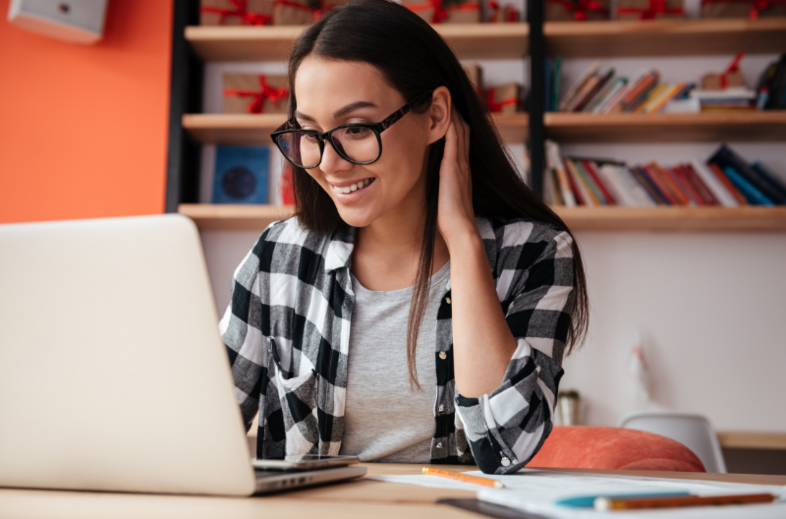 Ung kvinna med glasögon sitter vid ett skrivbord och arbetar på en bärbar dator i ett hemmakontor.