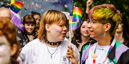 Två personer håller regnbågsflaggor på en prideparad, omgivna av bubblor och andra deltagare.