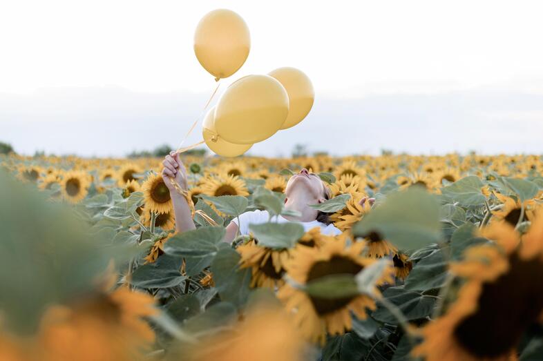 En person håller tre gula ballonger i ett fält av solrosor under en ljus himmel.