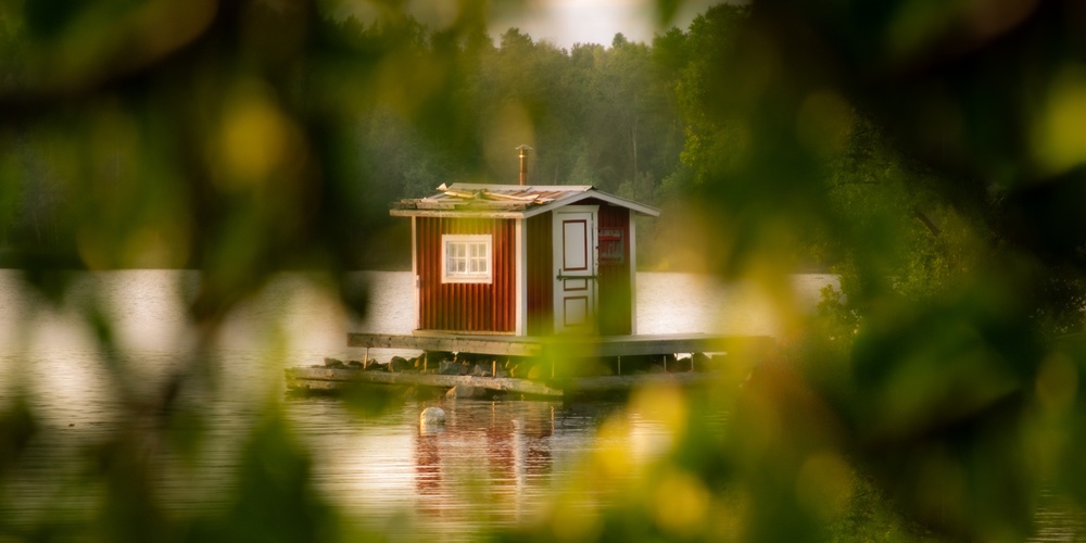 Ett litet rött hus på en brygga vid en sjö, omgiven av suddiga gröna trädgrenar i förgrunden.