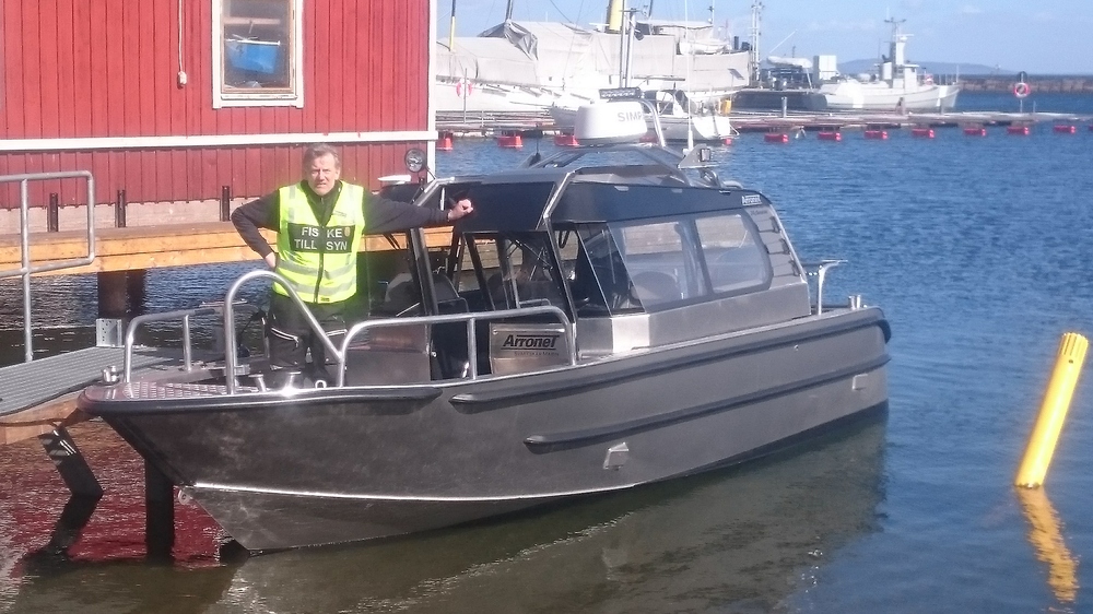 Person i gul väst står på en aluminiumbåt vid en brygga, med röda byggnader och båtar i bakgrunden.