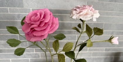 Två konstgjorda rosor av papper, en rosa och en ljusrosa, står mot en ljusgrå kakelvägg.