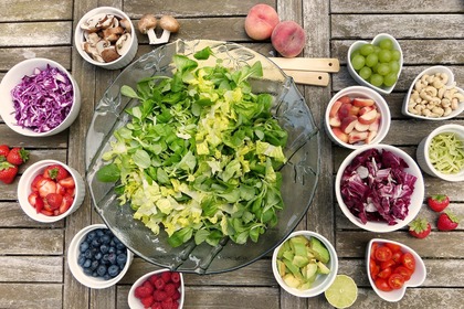 En stor salladsskål med grönsaker omgiven av små skålar med frukt, bär, nötter och grönsaker på ett träbord.