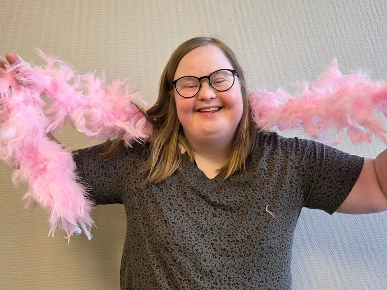 Person med axellångt hår håller en rosa fjäderboa runt nacken mot en ljus vägg.