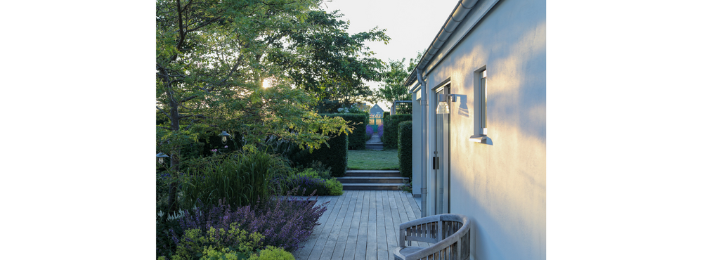 Trädgård med trädäck, grönska och modern husfasad i kvällssol.