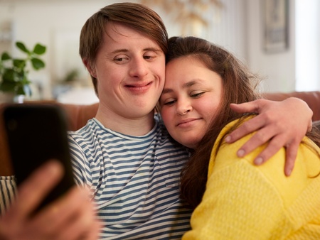 Två unga personer sitter i en soffa och tar en selfie tillsammans, de kramar varandra och ser glada ut.