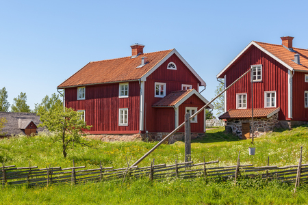 Två röda trähus med vita knutar på landet, omgivna av gräs, staket och blå himmel.