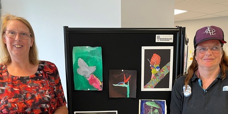 Två kvinnor står bredvid en svart utställningstavla med färgglada teckningar i ett ljust rum.