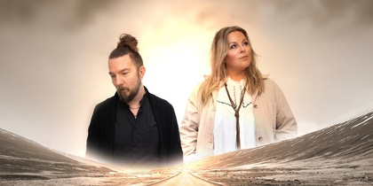 Två personer står på en öde väg i ett öppet landskap med dimmig himmel.