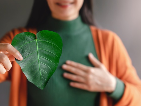 Kvinna håller ett grönt hjärtformat blad framför bröstet, klädd i grön tröja och orange kofta.