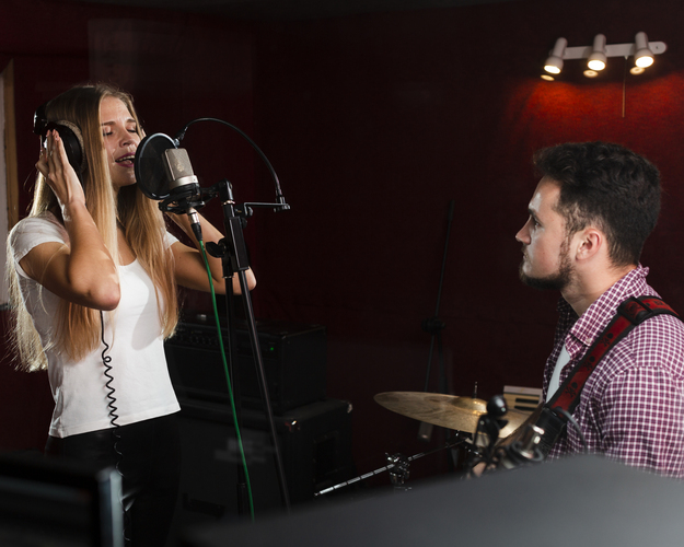 En kvinna sjunger i mikrofon med hörlurar, en man spelar gitarr i en musikstudio.