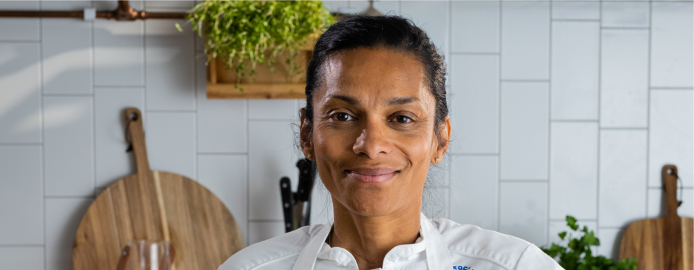 Person i vit kockrock står i ett kök med vita kakelväggar och träskärbrädor i bakgrunden.