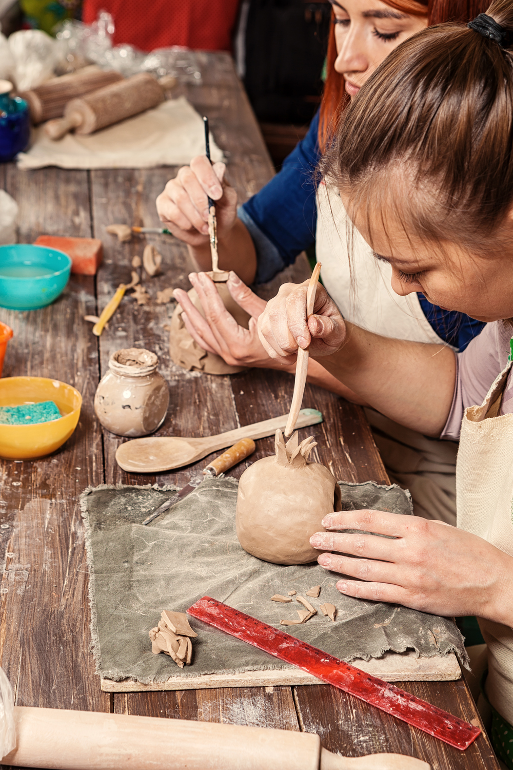 Två personer arbetar med keramik på ett träbord, formar lera med verktyg och händer.