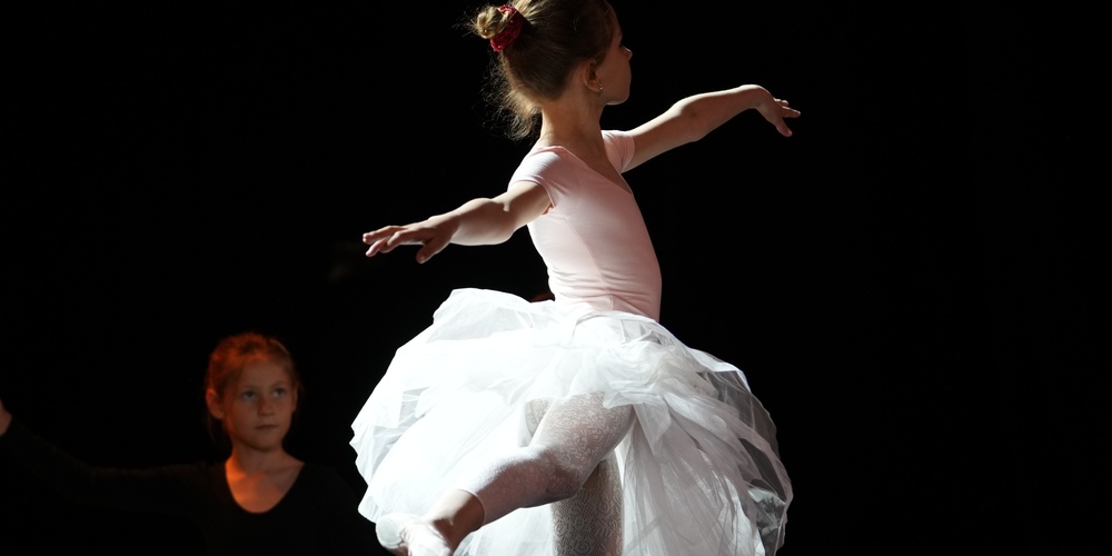 En ung ballerina i vit tyllkjol dansar på scenen med armarna utsträckta, belyst av scenljus.