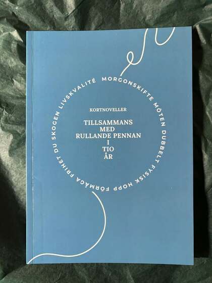 Blå bok med vit text på omslaget, liggande på ett grönt tyg. Titeln är 'Tillsammans med rullande pennan i tio år'.