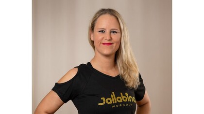 Person med långt blont hår och svart t-shirt med texten 'Jalabina Workout' står mot en neutral bakgrund.
