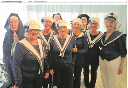 Grupp äldre kvinnor klädda i sjömanskläder och några i mörka dräkter, står tillsammans inomhus.