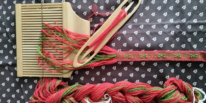 Vävredskap, garn och ett vävt band i rött och grönt på ett mönstrat tyg.