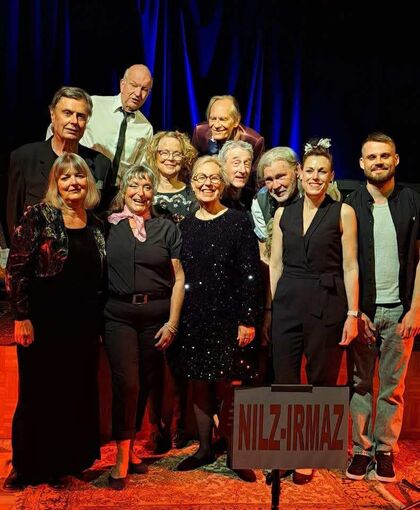 En grupp människor står tillsammans på en scen med mörk bakgrund och ett skylt med texten 'NILZ-IRMAZ' framför dem.
