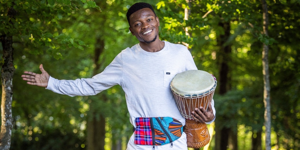 En leende man står barfota på en trästig i skogen och håller en trumma under armen.
