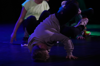 En person utför ett breakdance-move på golvet med stöd av ena handen, andra personer i bakgrunden.