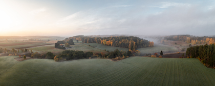 Panoramabild över ett dimmigt landskap med åkrar, skog och morgonljus.