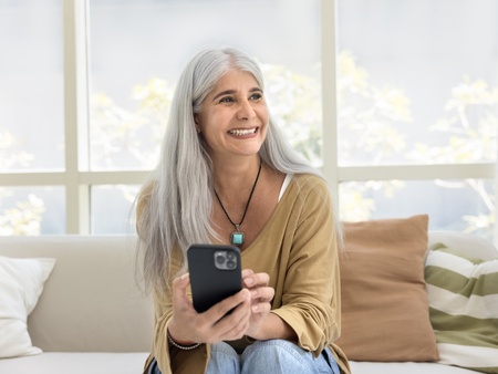 Person med långt grått hår sitter i soffa och håller en mobiltelefon i vardagsrum med stora fönster.