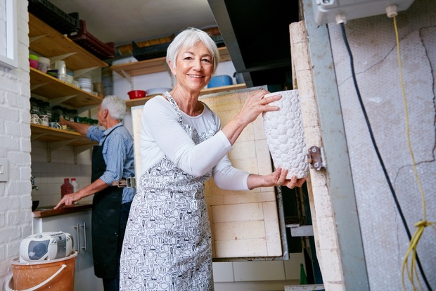 Äldre kvinna öppnar en keramikugn i en ljus verkstad, en man arbetar i bakgrunden.