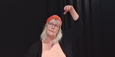 Kvinna med glasögon och röd sjal på huvudet sitter på knä och gestikulerar på en scen med tända ljus och tyg framför sig.