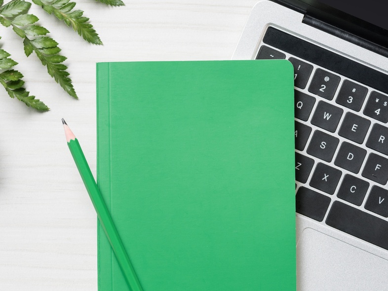 Grön anteckningsbok och penna på en bärbar dator, med växt och ormbunksblad på ett vitt skrivbord.