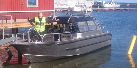 En person i gul väst står på en metallbåt vid en brygga med röda byggnader och båtar i bakgrunden.