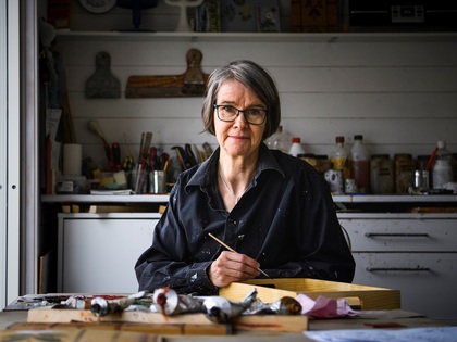 Person med glasögon och mörk skjorta sitter vid ett bord i en ateljé och målar med pensel.