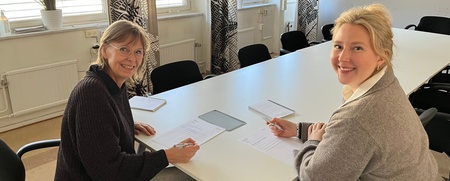 Två kvinnor sitter vid ett konferensbord och skriver på papper, båda ler mot kameran.