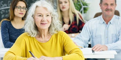 Äldre kvinna i gul tröja sitter i klassrum med andra vuxna och antecknar.