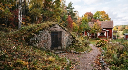 En gammal jordkällare av sten och ett rött trähus omgivna av höstfärgad skog och trädgård.