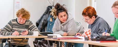 Fyra personer sitter vid ett bord och arbetar tillsammans med papper och pennor i ett klassrum.
