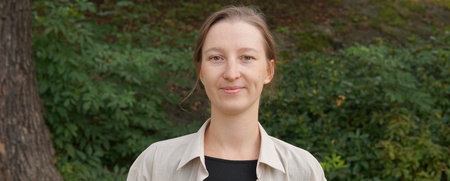 Kvinna med ljust hår står utomhus framför grönska och ler mot kameran.