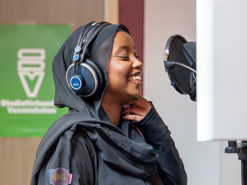 Kvinna med hijab och hörlurar sjunger i en mikrofon i en inspelningsstudio.