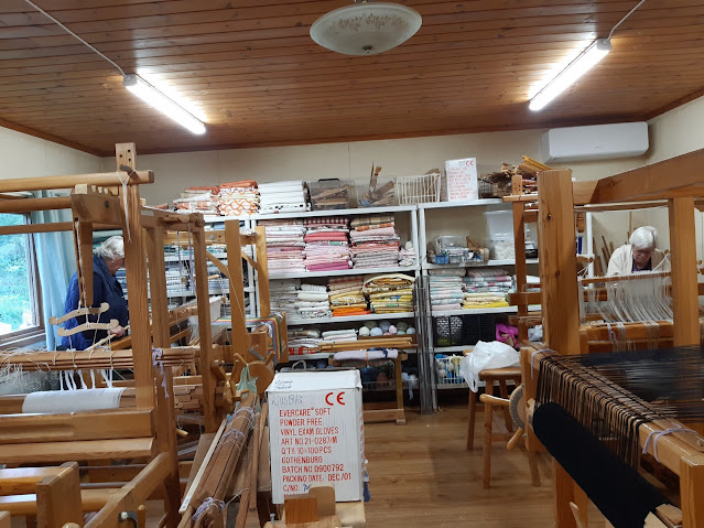 Vävstuga med flera vävstolar, hyllor med tyg och två personer som arbetar vid vävstolarna.