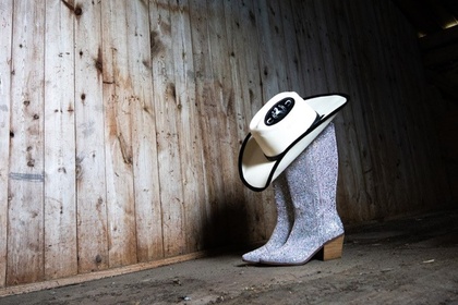 Ett par glittriga cowboyboots med en cowboyhatt ovanpå står på ett trägolv i en gammal lada.