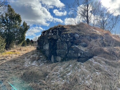En gammal stenkällare täckt av gräs och mossa i ett öppet landskap under en blå himmel med moln.