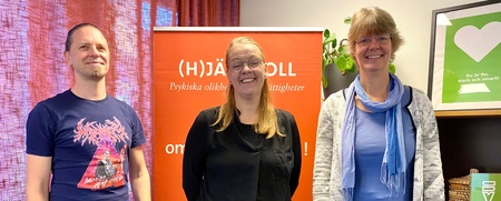 Tre personer står framför en orange skylt inomhus, leende mot kameran.