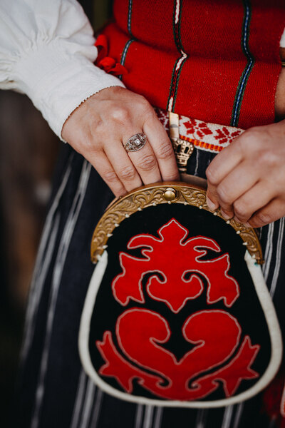 Närbild av händer som håller en traditionell väska med röd och svart brodyr, del av folkdräkt.