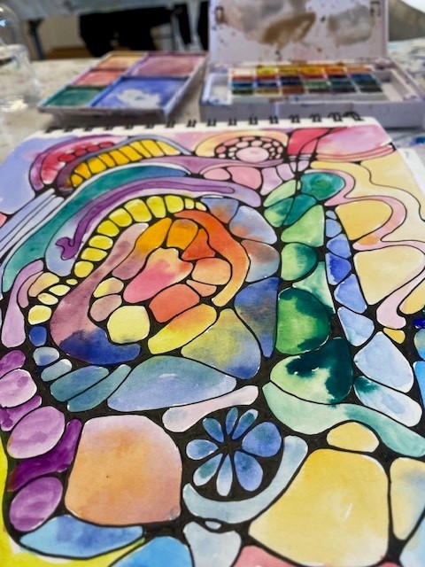 Abstrakt akvarellmålning med färgglada former och svarta konturer, målarlåda syns i bakgrunden.