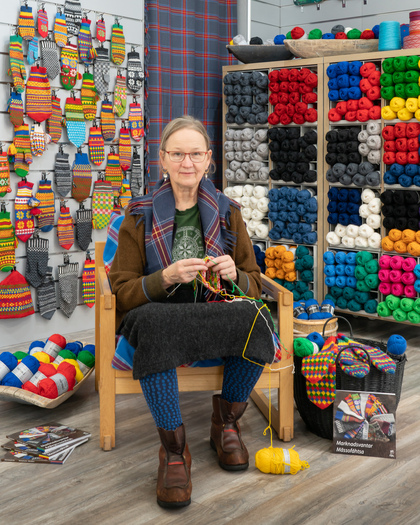 En kvinna sitter och stickar i en garnbutik omgiven av färgglada garnnystan och stickade vantar på väggen.