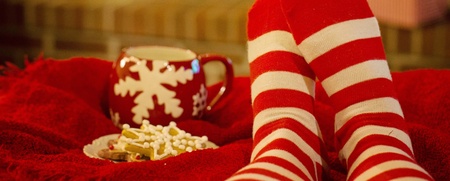 Person med röda och vita randiga strumpor vilar framför en öppen spis, med en julmugg och kakor på en filt.